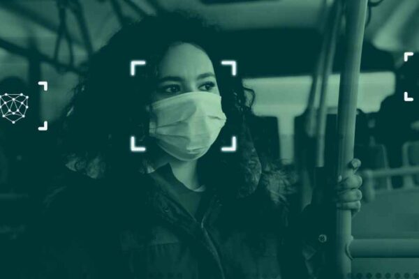 A person with a facemask on a bus with two others in the back. Each seem to be digitally tracked by facial recognition techonolgy that reads points on their face.