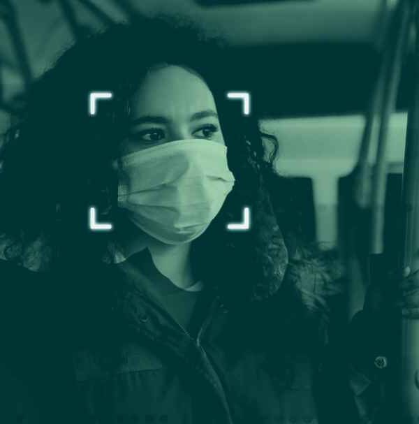 A person with a facemask on a bus with two others in the back. Each seem to be digitally tracked by facial recognition techonolgy that reads points on their face.