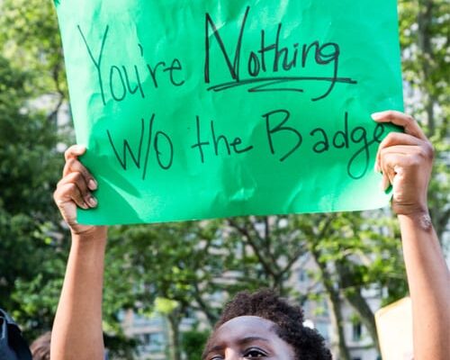 A masked person raises a green posterboard wtih the words "You're Nothing W/O the Badge"