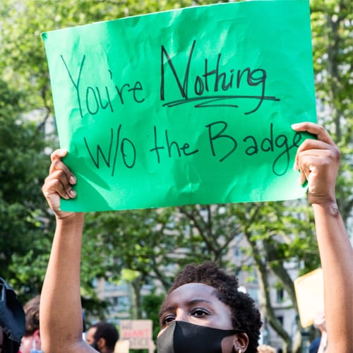 A masked person raises a green posterboard wtih the words "You're Nothing W/O the Badge"