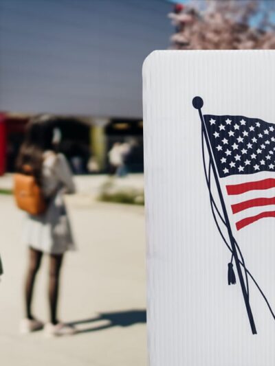 A person with a mask on holds a clutch wallet and is standing in front of a plastic voting divider with a print of the American flag and the word VOTE.
