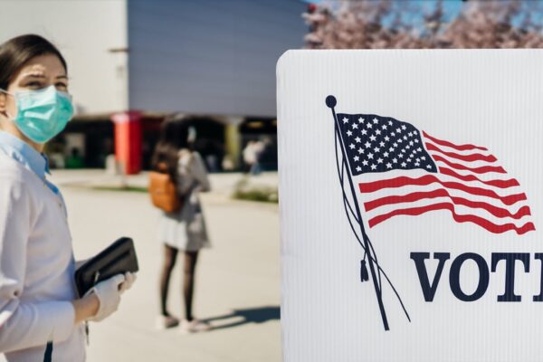 A person with a mask on holds a clutch wallet and is standing in front of a plastic voting divider with a print of the American flag and the word VOTE.