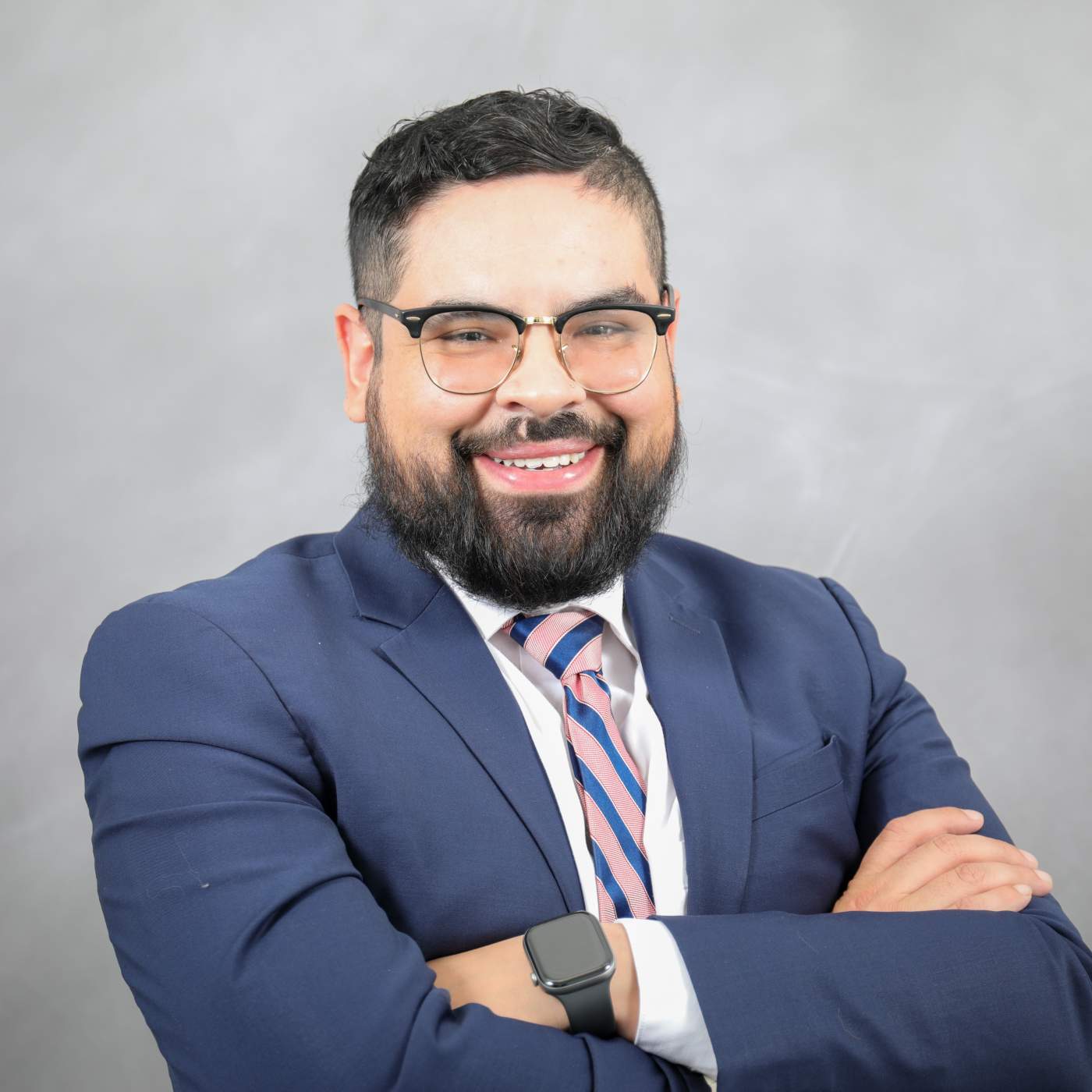 Photo of Andres (man with dark hair, glasses, dark beard, wearing a navy suit with a pink and navy tie) smiling with his arms crossed.