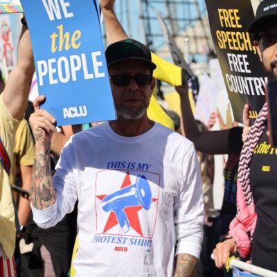 Person standing in a white long sleeve shirt that reads "This is my protest shirt." Also holding a blue sign that reads "We the People." They are wearing a hat and sunglasses.