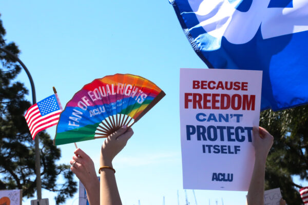 Hand holding rainbow fan that says "Fan of Equal Rights". Another sign says "Because Freedom Can't Protect Itself".