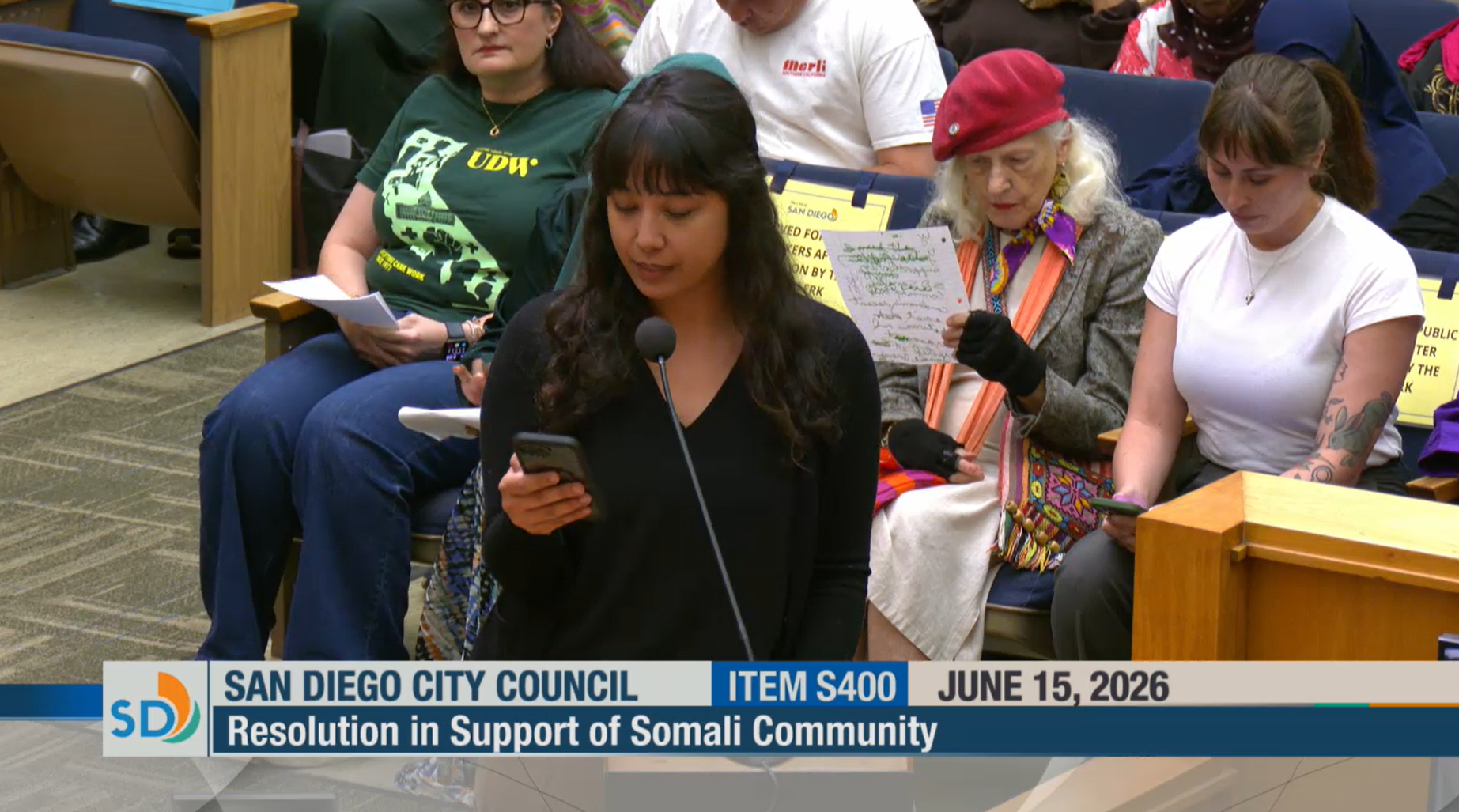 Samantha Itazawa (long dark hair, bangs, wearing black longsleeve shirt) delivers testimony into microphone standing before a seated crowd.
