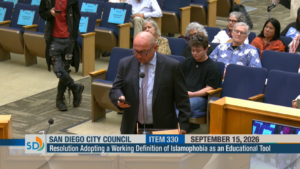 Eddie Meyer, Senior Policy Advocate at the ACLU-SDIC, gives public testimony at the San Diego City Council meeting. He is bald, wears black glasses, is wearing a black suit with a blue collared shirt and a purple tie. At the bottom of the image is a running header, that says "San Diego City Council; Item 330; September 15, 2026". Underneath is another bar that says "Resolution Adopting a Working Definition of Islamophobia as an Educational Tool".