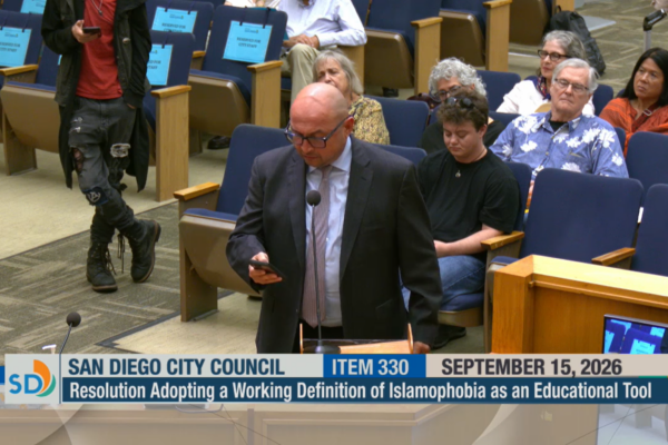 Eddie Meyer, Senior Policy Advocate at the ACLU-SDIC, gives public testimony at the San Diego City Council meeting. He is bald, wears black glasses, is wearing a black suit with a blue collared shirt and a purple tie. At the bottom of the image is a running header, that says "San Diego City Council; Item 330; September 15, 2026". Underneath is another bar that says "Resolution Adopting a Working Definition of Islamophobia as an Educational Tool".