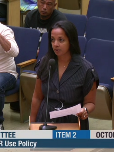 nalini gupta wears a black button up dress with a collar and has long black straight hair. She stands in front of a lectern with a microphone.