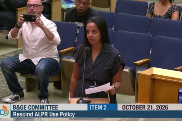 nalini gupta wears a black button up dress with a collar and has long black straight hair. She stands in front of a lectern with a microphone.