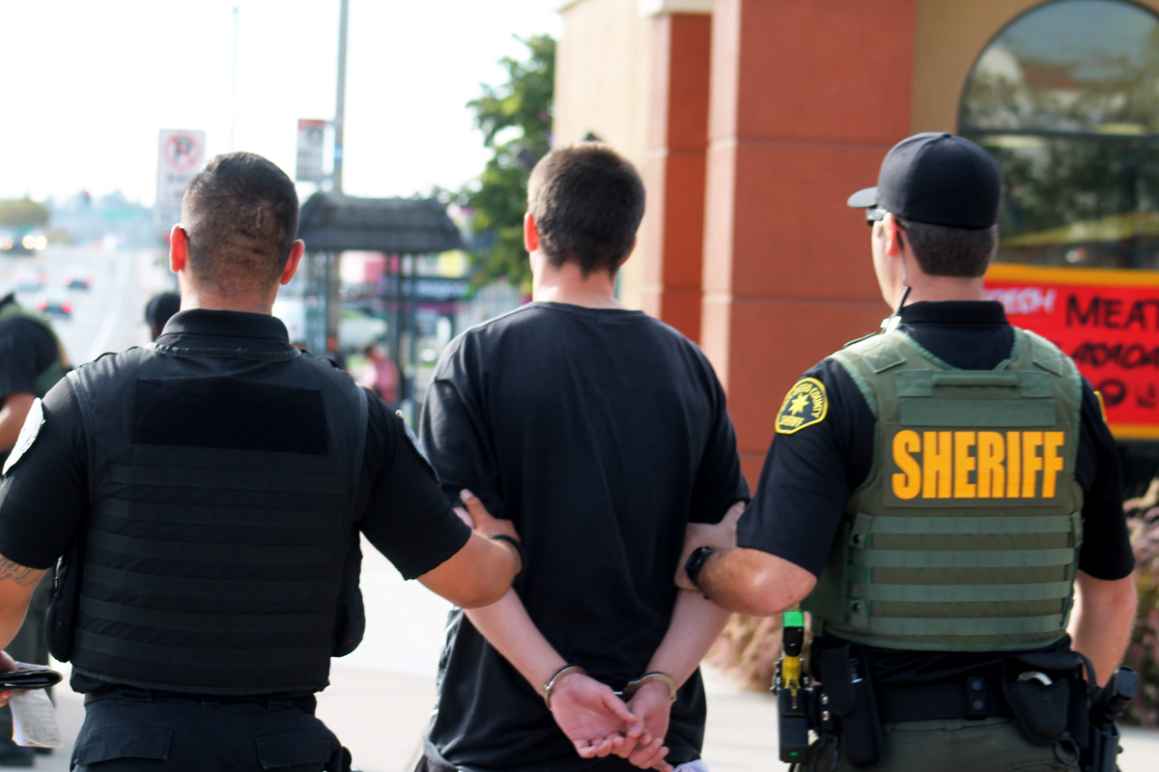 A male in handcuffs is being escorted away by a San Diego Police Department officer and a San Diego County Sherrif