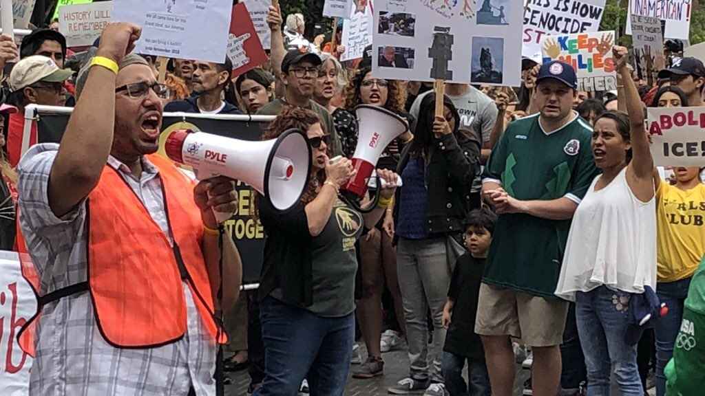 Man with glasses and a orange fluorescent safety vest has a raised fist and is yelling into a megaphone among a crowd of protestors