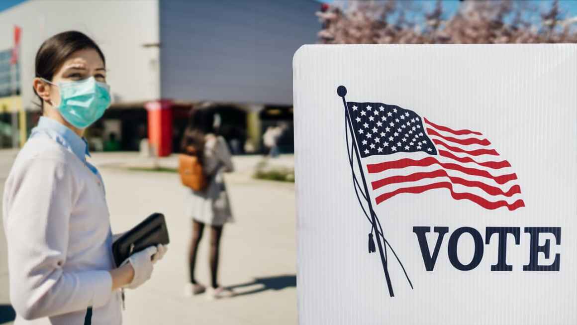 A person with a mask on holds a clutch wallet and is standing in front of a plastic voting divider with a print of the American flag and the word VOTE.