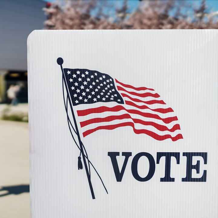 A person with a mask on holds a clutch wallet and is standing in front of a plastic voting divider with a print of the American flag and the word VOTE.